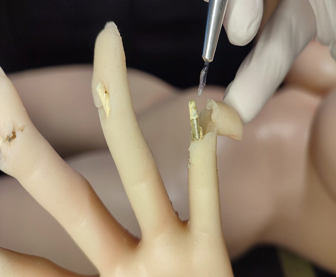A gloved hand using a tool to apply adhesive to a damaged finger of a mannequin-like doll, showing a repair process step.